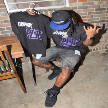 Model wearing Dinamik Tee Black/Purple with a second black tee on a hanger beside a brick wall.